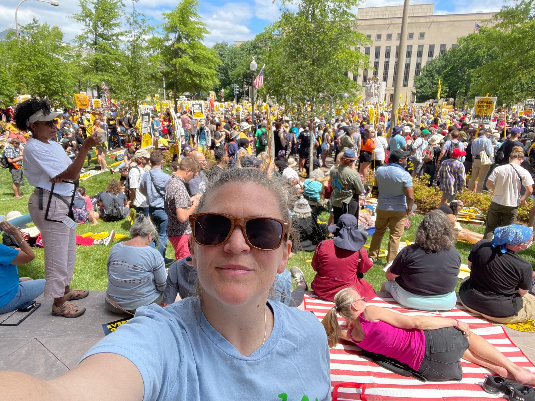 A person with sunglasses at a political rally.  In the background: people holding up signs