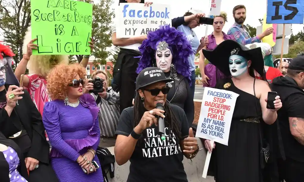 A Black person, speaking into a mic, wearing a T-shirt saying "Black names matter" and holding sign saying "Facebook: stop badering people for ID.  My name is as real as yours".  In the background, drag queens dressed as nuns, and other protestors, some also holding signs