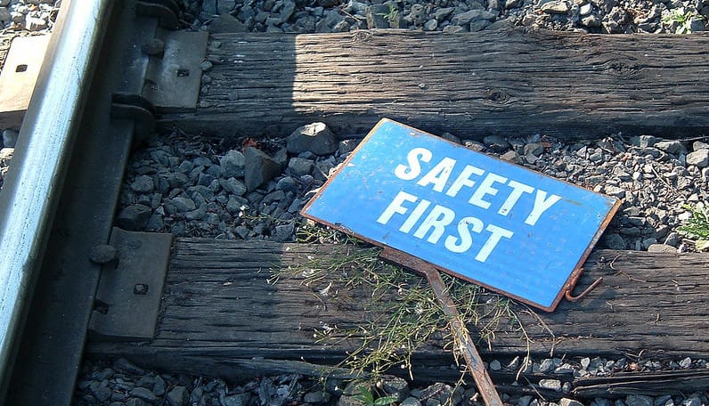 A closeup of railroad tracks, with a couple of wooden ties.  Lying on the ties, a blue sigh with white letters saying "Safety first"