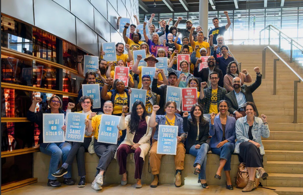 30 people sitting on stairs at Seattle City Hall, with hands raised, many with signs sayin things like "No AI on Aisle 5"
